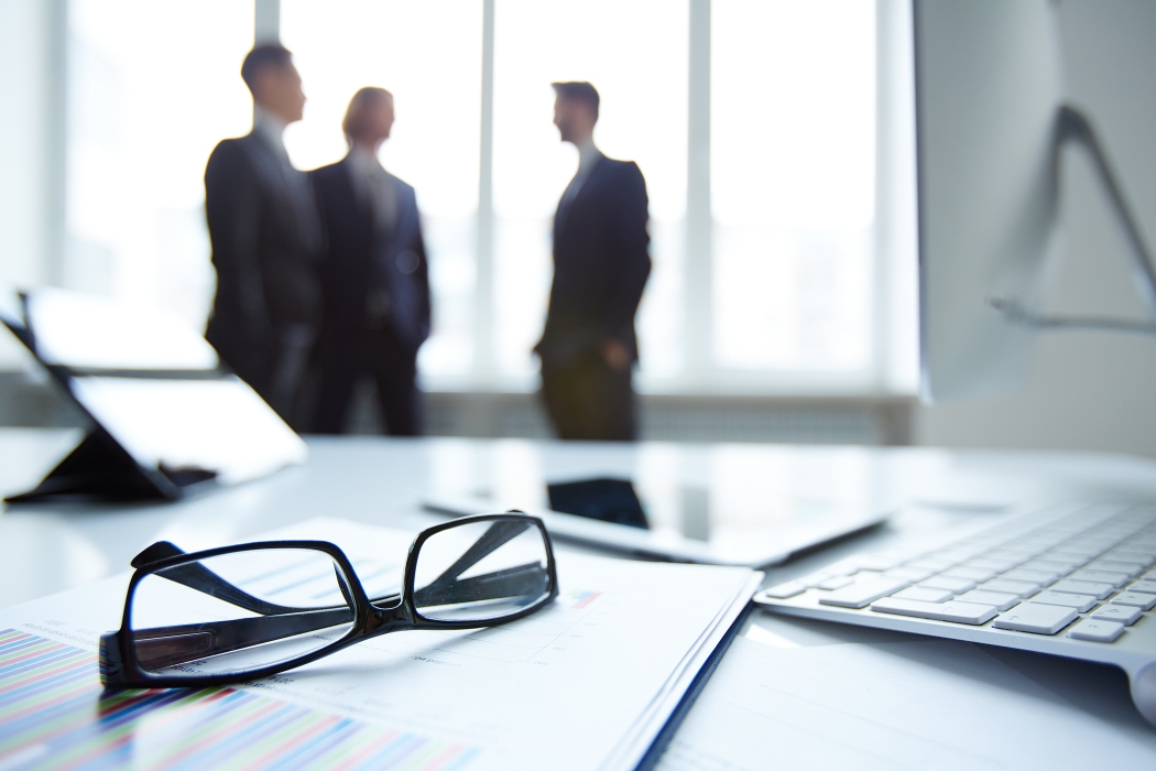 Technological devices, eyeglasses and financial document at workplace on background of three businessmen discussing ideas